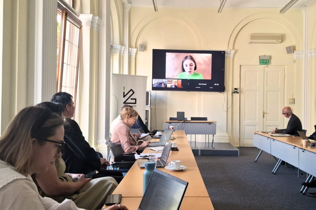 participants in a panel discussion sitting at desks set up in an oval shape. An online participant is visible on a large screen.